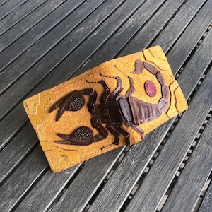 Pode incluir: Uma carteira de couro bege com um design de escorpião em relevo em marrom escuro. O escorpião tem garras texturizadas e uma cauda segmentada, com um pequeno detalhe circular vermelho. A carteira é retangular e parece ser feita à mão.
