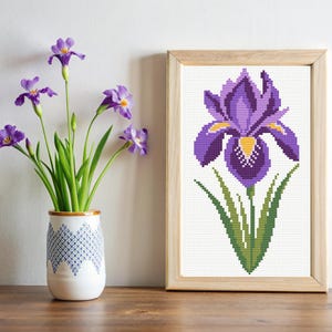 May include: A cross-stitch pattern of a purple iris flower with yellow accents, framed in a natural wood frame.