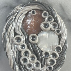 May include: A silver and white oval pendant with a brown, white, and silver swirl design. The pendant is decorated with small, round, silver beads.