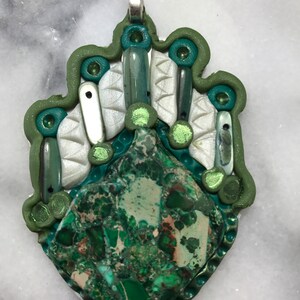 May include: A green and white pendant with a green stone in the center. The pendant is made of polymer clay and has a silver bail.