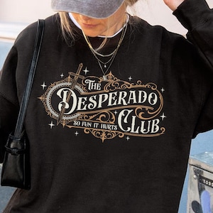 May include: Black sweatshirt with the text "The Desperado Club" in a decorative design. The design includes a sword and the phrase "So Fun It Hurts". The sweatshirt is worn with a baseball cap and a small black purse.