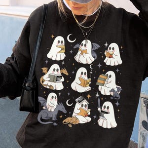 May include: Black sweatshirt with a pattern of white ghost illustrations reading books, with bats, dragons, and crescent moons. The design includes stars and small details, creating a whimsical and spooky aesthetic.