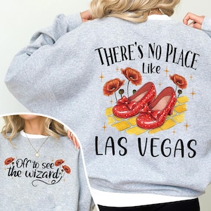 May include: A light gray sweatshirt with the text "THERE'S NO PLACE Like LAS VEGAS" and an illustration of red shoes, poppies, and a yellow brick road. The front of the sweatshirt has the text "Off to see the wizard".