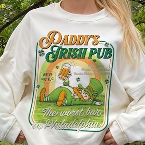 May include: A white crewneck sweatshirt featuring a graphic print that reads "Paddy's Irish Pub" and "The worst bar in Philadelphia." The design includes a cartoon leprechaun holding a beer mug, a rainbow, shamrocks, and the text "We've got egg!"