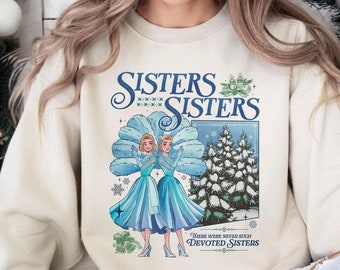 Puede incluir: Una sudadera beige de cuello redondo con un estampado gráfico de dos mujeres con vestidos azules y grandes abanicos de plumas, un pino nevado y el texto "SISTERS SISTERS" y "THERE WERE NEVER SUCH DEVOTED SISTERS" en tinta azul y verde.