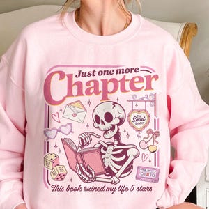 May include: A light pink crewneck sweatshirt featuring a graphic of a skeleton reading a book. The text reads Just one more Chapter, The Smut Reader, and This book ruined my life 5 stars. Additional illustrations include an envelope, dice, cherries, and a cassette tape.