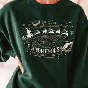 May include: Dark green sweatshirt with a graphic design featuring a sleigh, reindeer, and the text "Fly You Fools." The design includes a ring, snowflakes, and a feather quill. The sweatshirt is a crew neck style.