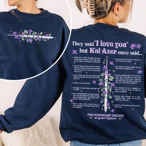 May include: A navy blue sweatshirt with a quote from the book "The Powerless Trilogy" by Lauren Roberts. The quote reads "They said "I love you" but Kai Azer once said..." and includes a list of 12 points. The sweatshirt features a purple floral design with a sword.