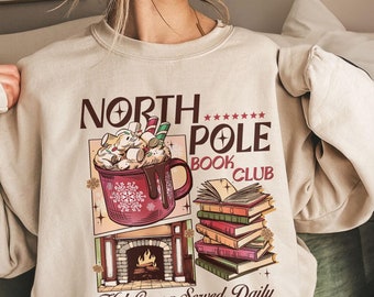 May include: A beige crewneck sweatshirt featuring a graphic print with the text North Pole Book Club and Hot Cocoa Served Daily. The design includes a red mug of hot cocoa with marshmallows, a stack of colorful books, and a fireplace illustration.