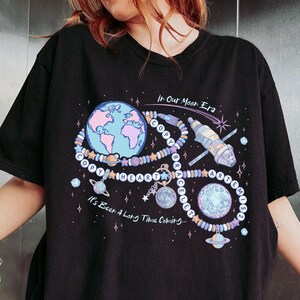 May include: A black t-shirt featuring a graphic of a beaded bracelet shaped like an infinity symbol, incorporating planets, a satellite, and the text "In Our Moon Era" and "It's Been A Long Time Coming" with "COPY HEART BRACELET ARTEMIS 2" on beads.