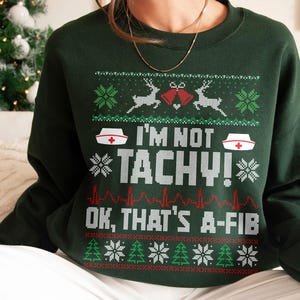 May include: Dark green sweatshirt with a festive design. The design includes reindeer, snowflakes, and the text "I'M NOT TACHY! OK, THAT'S A-FIB" in white and red. A fun, medical-themed holiday sweater.