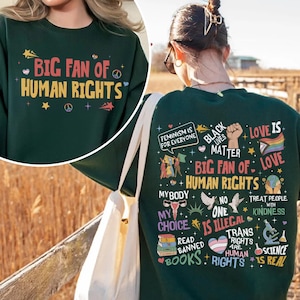 May include: A dark green long-sleeve sweatshirt featuring a graphic print. The front displays the text "BIG FAN OF HUMAN RIGHTS" with stars and peace signs. The back features various social justice slogans, icons, and the text "BIG FAN OF HUMAN RIGHTS".