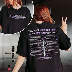 May include: Black t-shirt with a purple and white graphic of a sword and flowers. The text "They said 'I love you' but Kai Azer once said..." is printed on the shirt. The text is a list of 12 numbered points. The bottom of the shirt says "The Powerless Trilogy by Lauren Roberts".