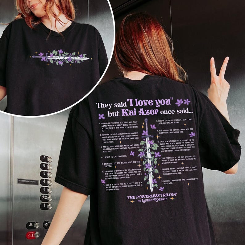 May include: Black t-shirt with a purple and white graphic of a sword and flowers. The text "They said 'I love you' but Kai Azer once said..." is printed on the shirt. The text is a list of 12 numbered points. The bottom of the shirt says "The Powerless Trilogy by Lauren Roberts".
