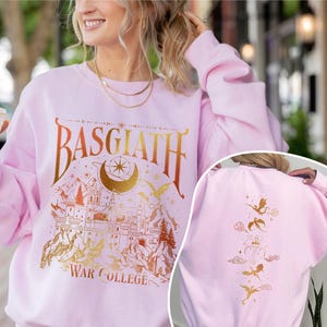 K&ouml;nnte beinhalten: Rosafarbener Pullover mit einem goldenen Grafikdruck eines Schlosses, Mondes, Sternen und Drachen. Der Text "Basgiath War College" ist auf der Vorderseite des Pullovers aufgedruckt.