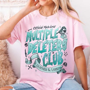 May include: Pink t-shirt with a green and blue graphic design that says "Official Member Multiple Deleters Club Leather & Lark". The graphic also includes a bird, a skull, a knife, and musical notes.
