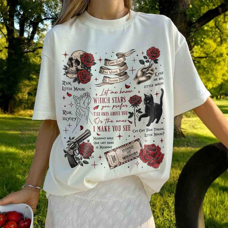 May include: White t-shirt with a black cat, red roses, and a skull graphic. The text on the shirt reads "Let me know which stars you prefer the ones above you or the ones I make you see".
