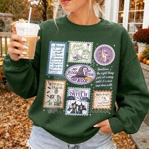 May include: A dark green long-sleeve sweatshirt featuring a graphic print of multiple postage stamp designs with witch-themed illustrations and text including "Witch's House," "My Blood Your Blood," and "There's a Little Witch in All of Us."