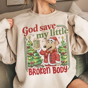May include: Beige sweatshirt with a Christmas-themed graphic. The graphic features a cartoon rat in a Santa hat and coat, holding a gift box. Text reads "God save my little BROKEN BODY" in red and green.