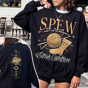May include: Black sweatshirt with gold text and graphics. The front features "S.P.E.W. Society for the Promotion of Elfish Welfare" with a yarn ball, knitting needles, and a sock. The back has a similar design with a yarn ball, needles, and celestial elements.