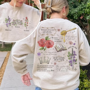 May include: A beige sweatshirt with a graphic print of flowers, crowns, swords, and text. The text includes quotes about love, power, and being a queen. The text reads: "Call us even call me crazy I don't care, just just call me yours. Oh darling I want to call you mine. For every girl who has ever felt powerless we are inevitable. We are tragedy. I would spend the rest of my days making her flower crowns if she wanted to be a queen. My queen, not kats, not ityas. Mine. You cannot be nothing when you are everything to someone else. Vicious, little thing."