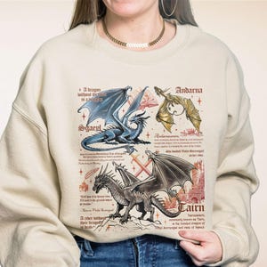 May include: A beige sweatshirt with a graphic print of three dragons and text about them. The text includes the names "Sgaepl", "Andarna", and "Cairn".