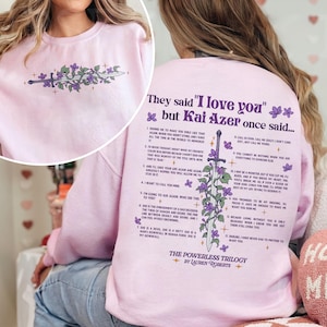 May include: Pink sweatshirt with a graphic print of a sword with purple flowers and the text "They said 'I love you' but Kai Azer once said..." and a list of numbered quotes. The text at the bottom of the graphic says "The Powerless Trilogy by Lauren Roberts".