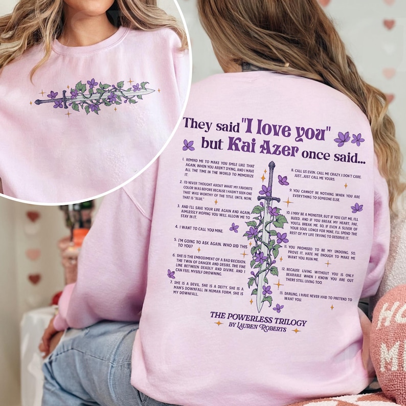 May include: Pink sweatshirt with a graphic print of a sword with purple flowers and the text "They said 'I love you' but Kai Azer once said..." and a list of numbered quotes. The text at the bottom of the graphic says "The Powerless Trilogy by Lauren Roberts".