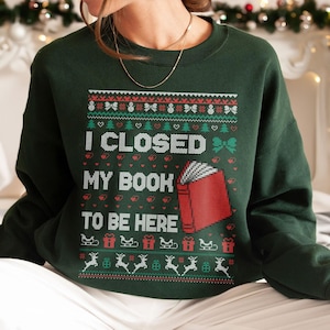 May include: A dark green crewneck sweatshirt featuring a festive knit-style graphic. The design includes the text I CLOSED MY BOOK TO BE HERE, a red book icon, and patterns of snowmen, bows, hearts, Christmas trees, sleighs, gifts, and reindeer.