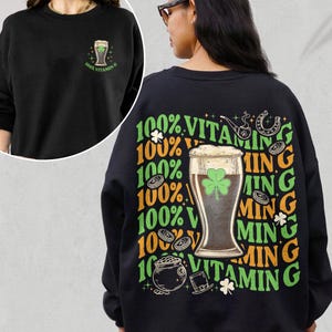 Puede incluir: Sudadera negra con tem&aacute;tica del D&iacute;a de San Patricio. La parte delantera presenta un peque&ntilde;o gr&aacute;fico de un vaso de cerveza con un tr&eacute;bol y el texto "100% Vitamin G". La parte trasera tiene un dise&ntilde;o m&aacute;s grande con un vaso de cerveza, tr&eacute;boles, monedas y el texto "100% Vitamin G".