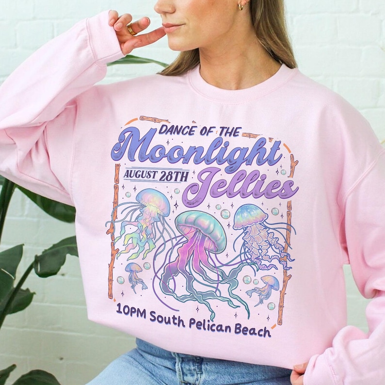 Puede incluir: Sudadera rosa claro con un dise&ntilde;o gr&aacute;fico. El dise&ntilde;o presenta el texto "Dance of the Moonlight Jellies", la fecha 28 de agosto e ilustraciones de medusas coloridas. Tambi&eacute;n se incluye la hora y el lugar del evento.