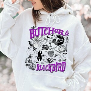 May include: White hoodie with a purple graphic that reads "Butcher & Blackbird." The graphic features various black and white illustrations, including a crow, spiderweb, skull, and chainsaw. The hoodie has a drawstring hood.