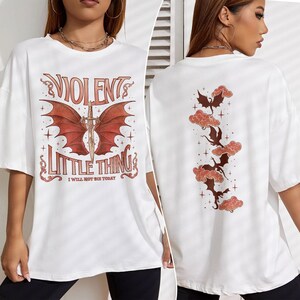 May include: White oversized t-shirt with a graphic print of a red dragon with wings and a sword. The text "VIOLENT LITTLE THING I WILL NOT DIE TODAY" is printed below the dragon. The back of the shirt has a graphic print of five red dragons flying in the clouds.