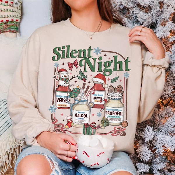 May include: A beige crewneck sweatshirt featuring the text Silent Night and a graphic illustration of four medicine bottles labeled Versed, Ativan, Haldol, and Propofol, decorated with Christmas elements like a snowman, Santa hat, and gifts.