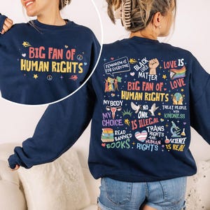 May include: A navy blue crewneck sweatshirt featuring a graphic print. The front displays the text "BIG FAN OF HUMAN RIGHTS". The back features a collage of colorful illustrations and text including "FEMINISM IS FOR EVERYONE", "BLACK LIVES MATTER", "MY BODY MY CHOICE", "READ BANNED BOOKS", and "SCIENCE IS REAL".