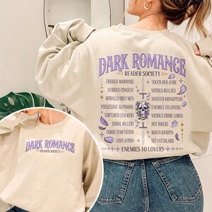 May include: A beige sweatshirt with the text "DARK ROMANCE READER SOCIETY" in purple. The back of the sweatshirt lists various romance novel tropes in a decorative layout, including "Trigger Warnings" and "Enemies to Lovers".