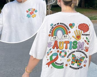 May include: A white t-shirt featuring a small colorful puzzle piece graphic on the front left chest. The back displays a large, vibrant design with a rainbow, a butterfly, a brain, a handprint, and the text "AUTISM IT'S OK TO BE DIFFERENT".