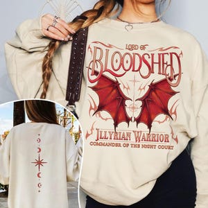 May include: A cream-colored sweatshirt with a red and black graphic print of a dragon with wings and the text "Lord of Bloodshed Illyrian Warrior Commander of the Night Court". The back of the sweatshirt has a red and white star and crescent moon design.
