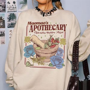 May include: Cream-colored sweatshirt featuring a graphic of a mortar and pestle with herbs and blue roses. The text reads "Maomao's Apothecary Remedies Medicine Poison" in a vintage style. A unique design for those interested in apothecary themes.