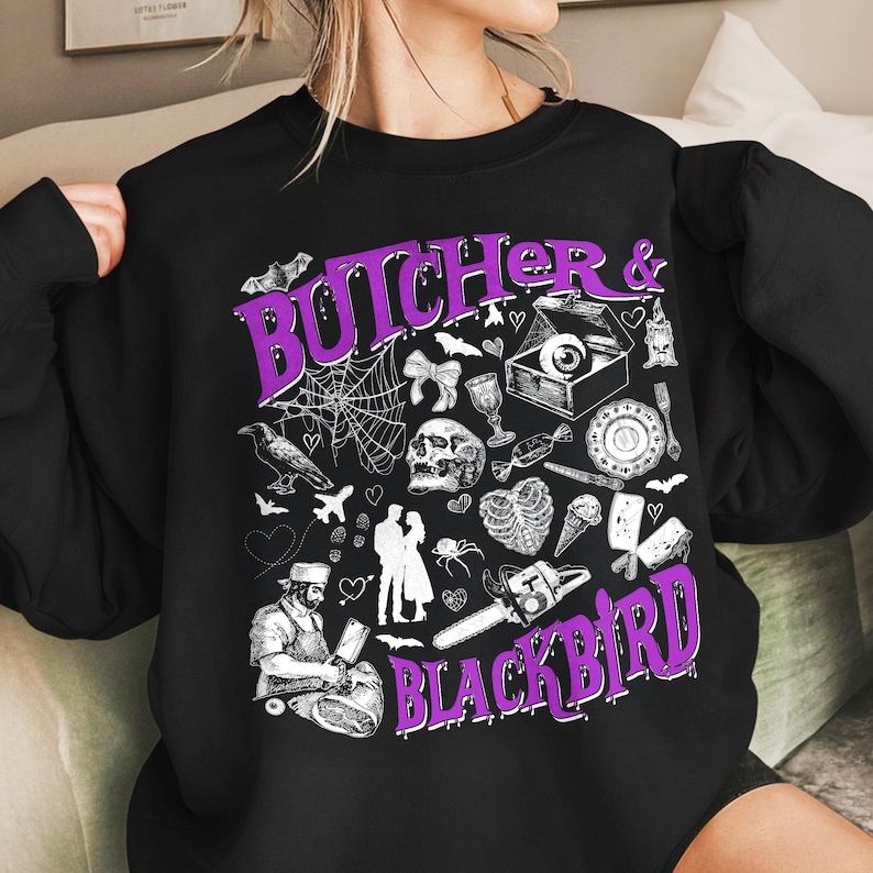 May include: Black sweatshirt with a purple graphic that reads "Butcher & Blackbird." The design includes a spiderweb, skull, chainsaw, and other spooky illustrations. The sweatshirt is a crew neck style.