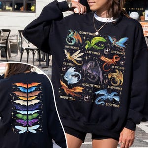 May include: A black long-sleeve sweatshirt featuring a graphic of various dragons labeled Rainwings, Hivewings, Leafwings, Silkwings, Icewings, Skywings, Nightwings, Sandwings, Mudwings, and Seawings. The back displays a vertical stack of colorful dragon wings.