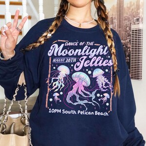 Puede incluir: Sudadera azul marino con un dise&ntilde;o gr&aacute;fico de medusas en tonos rosa, morado y azul. El texto dice "Dance of the Moonlight Jellies, August 28th, 10PM South Pelican Beach."