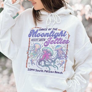 Puede incluir: Sudadera con capucha blanca con un gr&aacute;fico colorido de medusas y el texto "Dance of the Moonlight Jellies, August 28th, 10PM South Pelican Beach." El dise&ntilde;o incluye medusas moradas, rosas y azules.