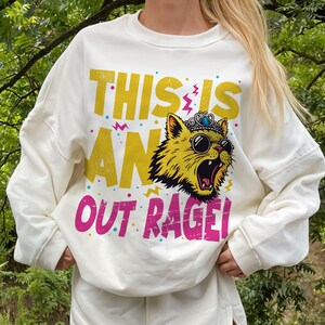 May include: Cream-colored sweatshirt with a graphic design. The design features a yellow cat wearing sunglasses and a crown, with the words "THIS IS AN OUT RAGE!" in yellow and pink text.
