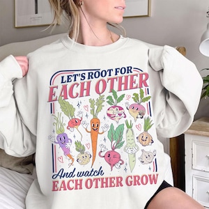 May include: Cream-colored sweatshirt with a colorful graphic of cartoon vegetables. The text reads "Let's Root For Each Other" and "And watch Each Other Grow". The design includes carrots, beets, and other root vegetables.
