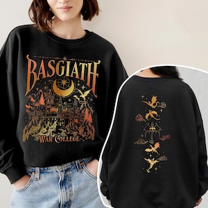 K&ouml;nnte beinhalten: Schwarzer Sweatshirt mit einem goldenen Grafikdruck eines Schlosses mit dem Text "Basgiath War College" und einem Halbmond mit Sternen. Die R&uuml;ckseite des Sweatshirts zeigt eine vertikale Reihe von f&uuml;nf goldenen Drachen-Silhouetten.