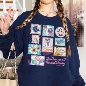 May include: Navy blue sweatshirt featuring a collage of colorful postage stamp designs. The stamps depict various images, including a laptop, a wedding suit, a woman, a sailboat, a bear, and the text "The Summer I Turned Pretty".