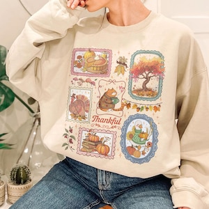 May include: A beige crewneck sweatshirt featuring a graphic print of various framed autumn-themed illustrations, including a pumpkin pie, a turkey, a cat holding a mug, a tree, and a goose, with the word Thankful written in cursive script.