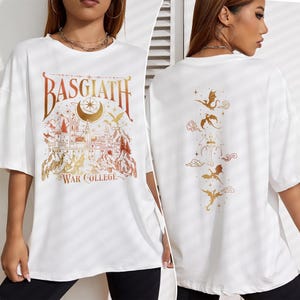K&ouml;nnte beinhalten: Wei&szlig;es Oversize-T-Shirt mit einem goldenen Grafikdruck eines Schlosses, Mondes, Sternen und Drachen. Der Text "Basgiath War College" ist auf der Vorderseite des Shirts gedruckt. Die R&uuml;ckseite des Shirts zeigt einen goldenen Grafikdruck von f&uuml;nf Drachen.