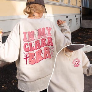 May include: A beige crewneck sweatshirt featuring a large pink graphic on the back with the text "IN MY CLARA ERA," a nutcracker, a ballerina, and small star and flower icons. A smaller matching circular graphic is printed on the front left chest.